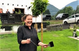 A woman stands laughing by an outdoor fireplace, surrounded by a green landscape and a building in the background.