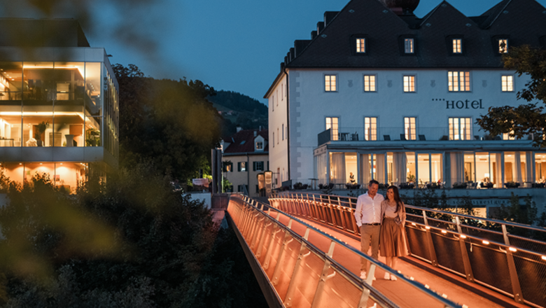 A couple walks across an illuminated bridge in front of a hotel in the evening.