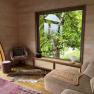 Innenansicht eines gem&uuml;tlichen Raumes mit gro&szlig;em Fenster, das auf einen gr&uuml;nen Baum blickt. Ein Sessel, ein Hocker und ein Sofa sind zu sehen.