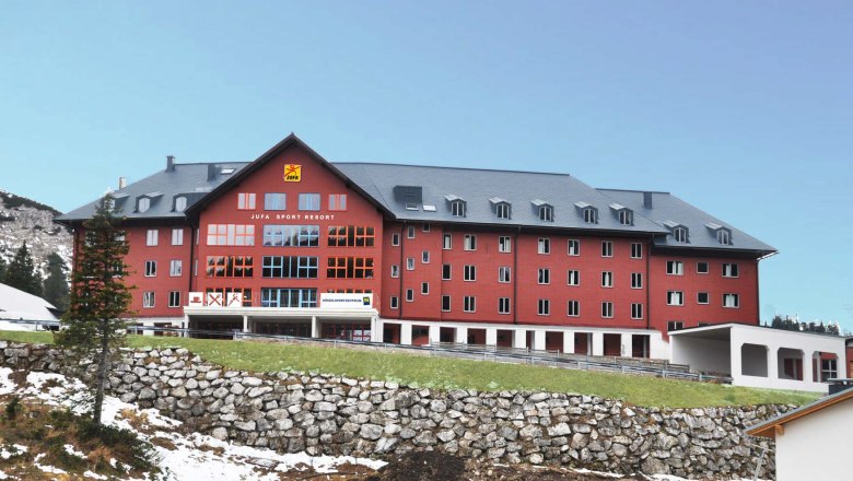 Großes rotes Gebäude des Jufa Sport Resorts in einer bergigen Landschaft.