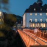 A couple walks across an illuminated bridge in front of a hotel in the evening.
