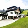 A traditional farmhouse with wood siding and flowers on the balcony, surrounded by green meadows and forests.