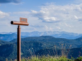 The majestic peaks of the Grosser &Ouml;tscher rise proudly against the clear sky, while the rolling hills and flowering meadows provide an inviting backdrop for hikers. A telescope invites you to enjoy the breathtaking view and experience the beauty of nature to the full.