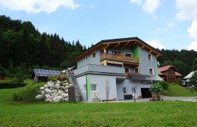 A modern house with wooden cladding and solar panels in a green, wooded setting.
