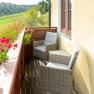Ein Balkon mit zwei geflochtenen St&uuml;hlen und einem Tisch, umgeben von bl&uuml;henden Blumen und mit Blick auf eine gr&uuml;ne Landschaft.