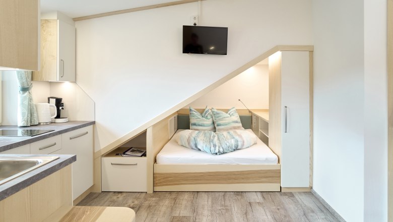 Kompaktes Apartment mit eingebautem Bett unter einer Dachschr&auml;ge, K&uuml;chenzeile und Fernseher.