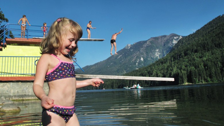 Children play and jump from a diving board into Lake Lunz, surrounded by mountains and trees.