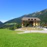 A yellow building with a balcony and flowers, surrounded by green meadows and mountains under a blue sky.