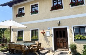 Ein traditionelles Bauernhaus mit gelber Fassade und roten Blumen in den Fenstern. Vor dem Haus steht ein Holztisch mit B&auml;nken und einem Sonnenschirm.