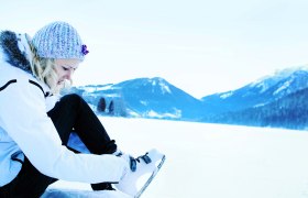 Person zieht Schlittschuhe an einem verschneiten See an, umgeben von Bergen.