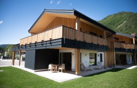Modern wooden architecture of a two-storey building with balcony and terrace, surrounded by green landscape.