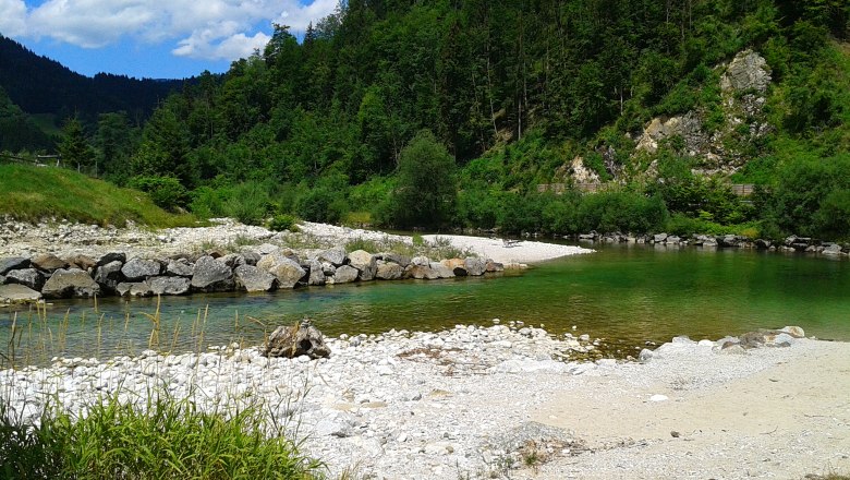 Flusslandschaft mit klarem Wasser, Kieselstrand und bewaldeten H&uuml;geln.