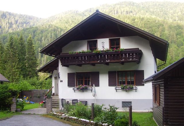 Ferienhaus Hager, © zVg Ein traditionelles Ferienhaus mit Holzbalkon und Blumenkästen, umgeben von grünen Bäumen und Bergen.