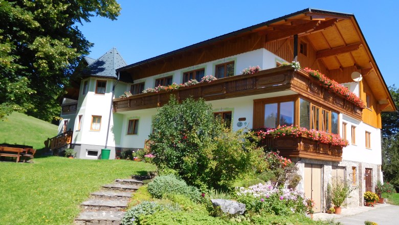 Bauernhof Köhr, © Bauernhof Köhr Ein traditionelles Bauernhaus mit Holzbalkon und blühenden Blumen, umgeben von grüner Landschaft und blauem Himmel.