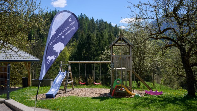 Spielplatz mit Rutsche und Schaukeln, umgeben von B&auml;umen und Natur.