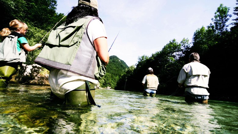 Gruppe von Menschen beim Fliegenfischen in einem Fluss, umgeben von Bäumen.