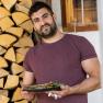 Ein Mann in einem weinroten T-Shirt h&auml;lt einen Teller mit Essen vor einem Stapel Holzscheite.