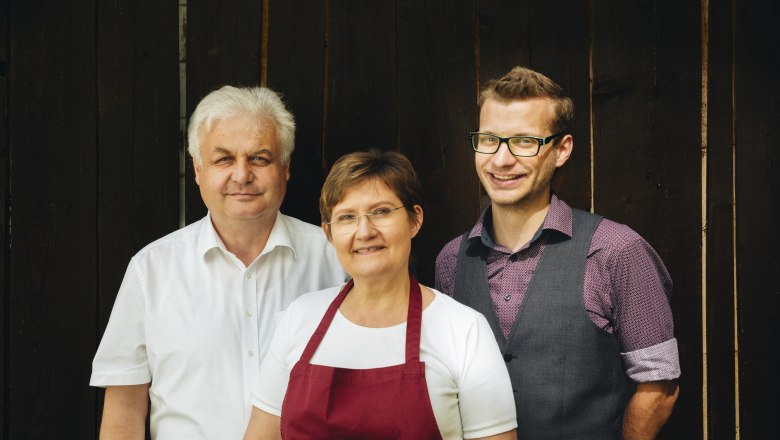 Three people stand in front of a wooden background, two men and a woman in the middle.