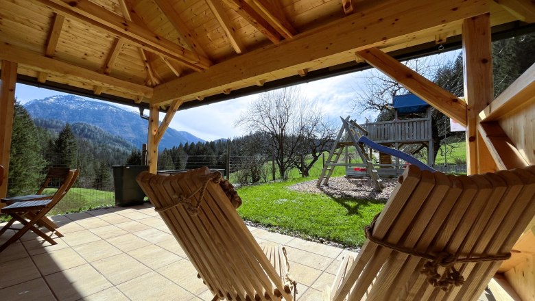 Blick aus einem Holzpavillon auf eine Berglandschaft mit Schaukel und Rutsche im Garten.