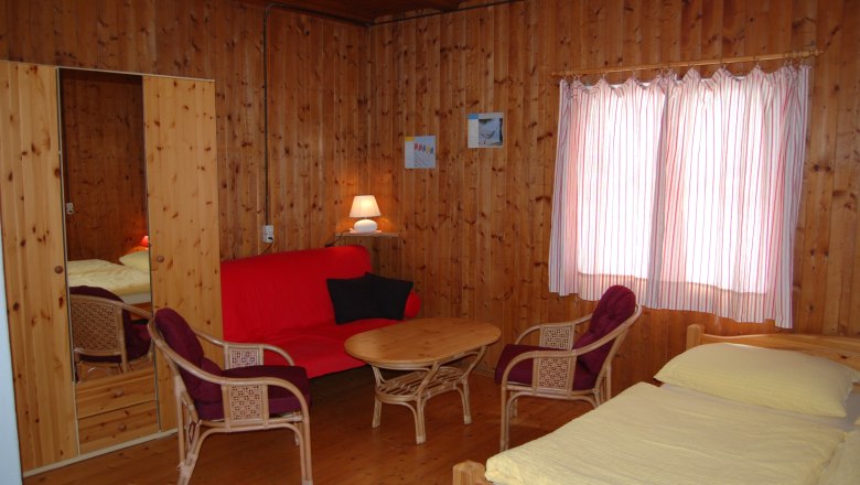 Innenansicht eines gemütlichen Zimmers mit Holzwänden, rotem Sofa, Tisch, Stühlen und Bett.