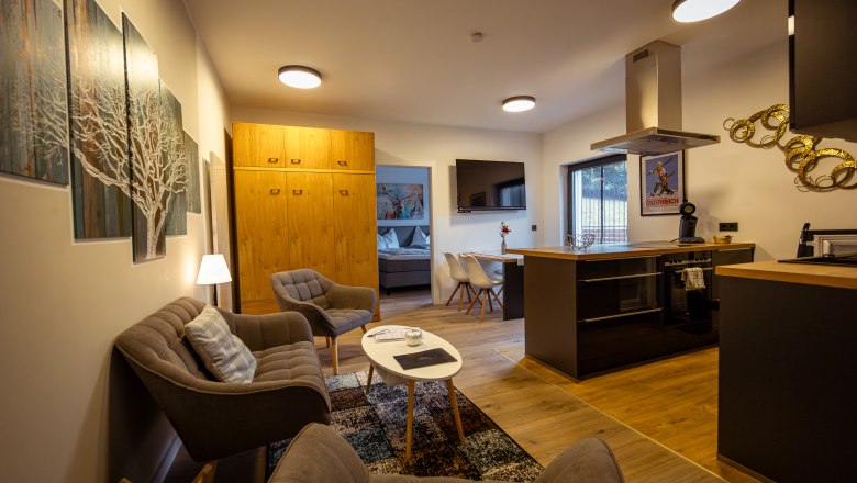 Modern living room with kitchen, sofa, armchairs and dining table. Light wood furniture and wall art can be seen.