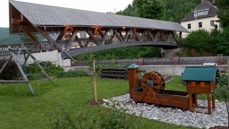 Überdachte Holzbrücke über einen Fluss mit einem kleinen Wasserrad im Vordergrund.