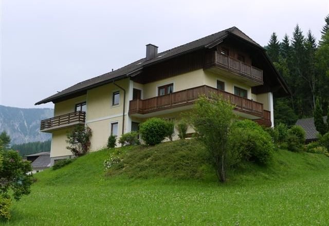 Ein zweistöckiges Haus mit Holzbalkonen auf einem grünen Hügel, umgeben von Bäumen und Wiesen.