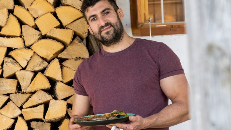 Ein Mann in einem weinroten T-Shirt h&auml;lt einen Teller mit Essen vor einem Stapel Holzscheite.