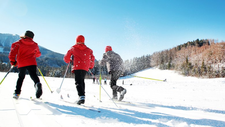 Three people cross-country skiing on a snow-covered trail under a blue sky.