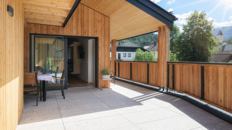 Moderne Holzterrasse mit Tisch und St&uuml;hlen, umgeben von B&auml;umen und Geb&auml;uden im Hintergrund.