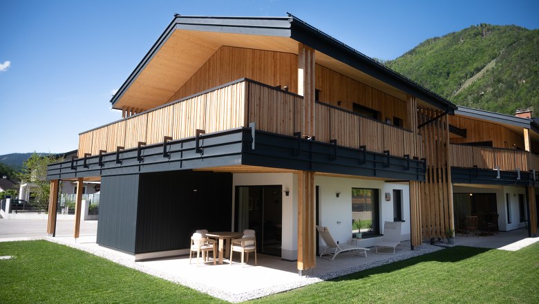 Moderne Holzarchitektur eines zweist&ouml;ckigen Geb&auml;udes mit Balkon und Terrasse, umgeben von gr&uuml;ner Landschaft.