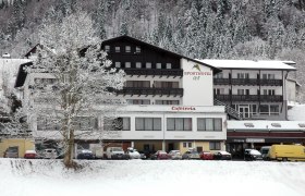 A snow-covered hotel building with the inscription 'Sporthotel' and 'Cafeteria', surrounded by snow-covered trees and parked cars.