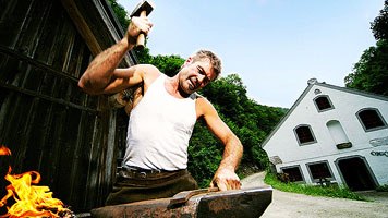 A blacksmith works with a hammer on an anvil in front of a traditional building.