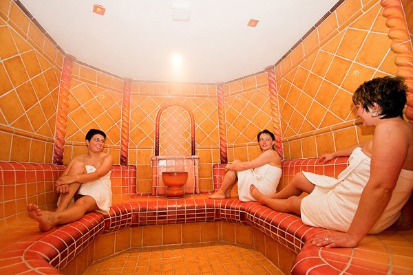 Drei Personen in einem gefliesten Dampfbad, eingewickelt in Handtücher.
