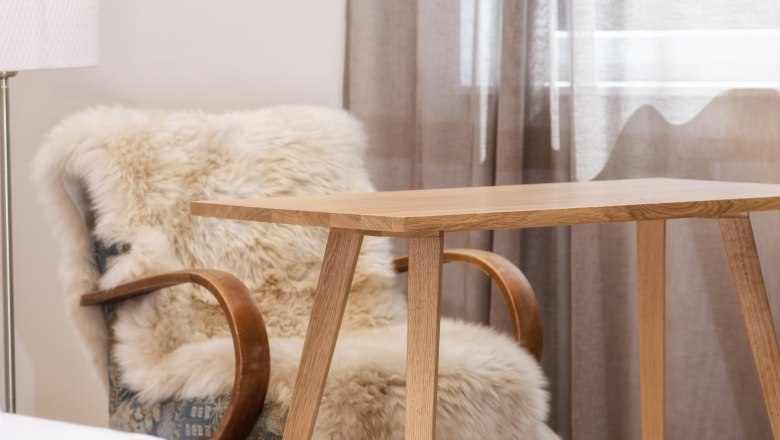 Ein gem&uuml;tlicher Raum mit einem Holztisch, einem Stuhl mit Fell&uuml;berwurf und einem Fenster mit Vorh&auml;ngen.