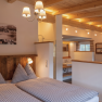 A cozy bedroom with a wooden beam ceiling and a double bed, decorated with checkered pillows and blankets. In the background is a bathroom with a sink; behind the half-height wall is a freestanding bathtub.