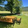 A garden with a wooden table and benches, surrounded by trees and meadows, with a playground in the background.