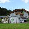 Ein modernes Haus mit Holzverkleidung und Solarpanelen in einer grünen, bewaldeten Umgebung.
