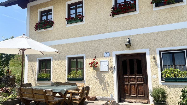 Ein traditionelles Bauernhaus mit gelber Fassade und roten Blumen in den Fenstern. Vor dem Haus steht ein Holztisch mit B&auml;nken und einem Sonnenschirm.