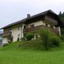 A two-storey house with wooden balconies on a green hill surrounded by trees and meadows.