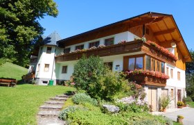 Ein traditionelles Bauernhaus mit Holzbalkon und bl&uuml;henden Blumen, umgeben von gr&uuml;ner Landschaft und blauem Himmel.