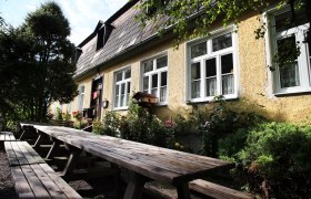 Gelbes Geb&auml;ude mit wei&szlig;en Fenstern und langen Holzb&auml;nken im Vordergrund.