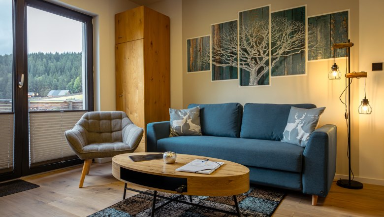 Cozy living room with blue couch, armchair, wooden table and forest view through the window.