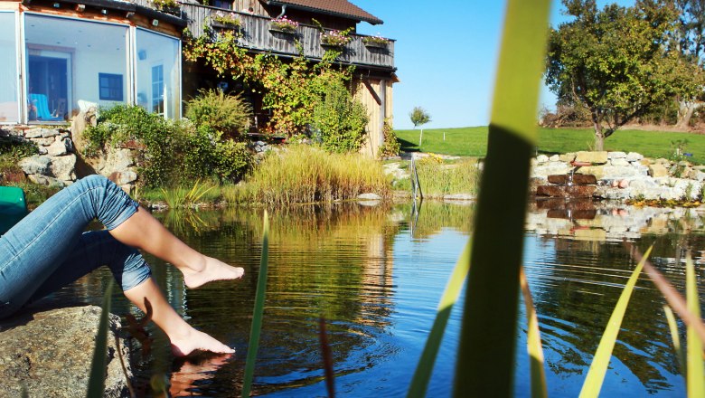 Person sitzt am Teichrand mit den Füßen im Wasser, vor einem Bauernhaus mit Garten.