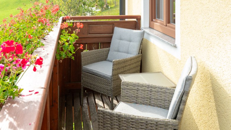 Ein Balkon mit zwei geflochtenen St&uuml;hlen und einem Tisch, umgeben von bl&uuml;henden Blumen und mit Blick auf eine gr&uuml;ne Landschaft.
