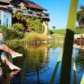 Person sitzt am Teichrand mit den Füßen im Wasser, vor einem Bauernhaus mit Garten.