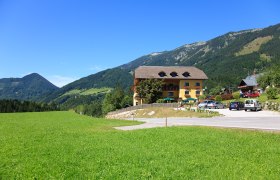 Ein gelbes Geb&auml;ude mit Balkon und Blumen, umgeben von gr&uuml;nen Wiesen und Bergen unter blauem Himmel.
